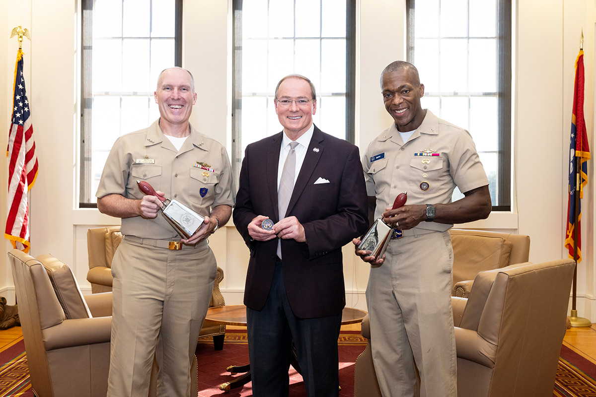 Two men in khaki navy uniforms holding cowbells flanking man in suit holding coin